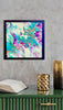 Carnival abstract painting in a black frame on a grey wall above a green cabinet with a book and gold decorative accents