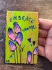 A hand holding a small rectangular handmade painting with scalloped edges, featuring the words 'EMBRACE change' in blue marker over a yellow-green background with pink and blue flower illustrations, placed on a dark wooden surface.