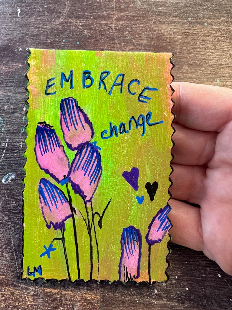 A hand holding a small rectangular handmade painting with scalloped edges, featuring the words 'EMBRACE change' in blue marker over a yellow-green background with pink and blue flower illustrations, placed on a dark wooden surface.