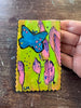 A hand holding a small, scalloped-edge handmade painting featuring a blue butterfly and pink stylized flowers on a bright yellow-green background, set against a dark wooden tabletop.