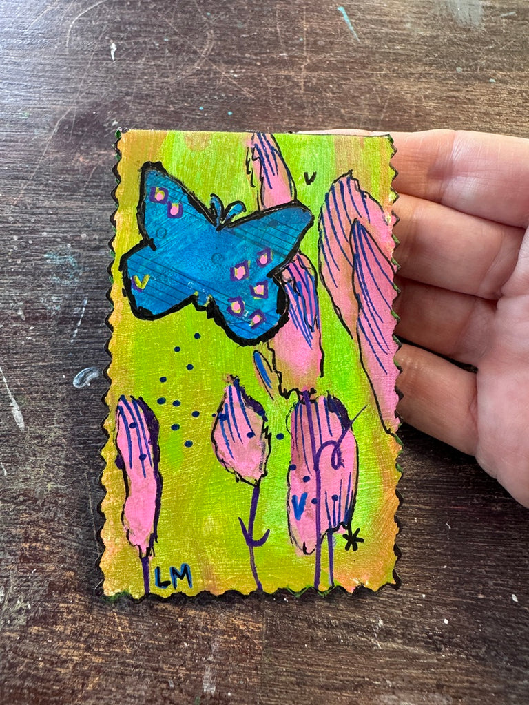 A hand holding a small, scalloped-edge handmade painting featuring a blue butterfly and pink stylized flowers on a bright yellow-green background, set against a dark wooden tabletop.