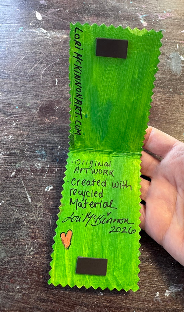 A hand holding a handmade green card or art print with serrated edges, handwritten text, two black rectangular stickers, and a small orange heart, photographed over a dark wooden surface.