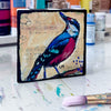a colorful bird art block painting on a table in front of small bottles of paint and a paint brush laying in front of it