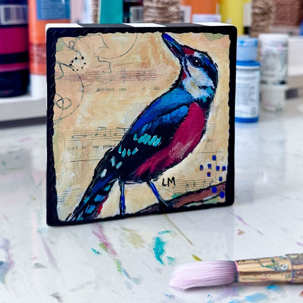 a colorful bird art block painting on a table in front of small bottles of paint and a paint brush laying in front of it