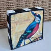a colorful bird art block painting on a white table with part of a basket showing behind it