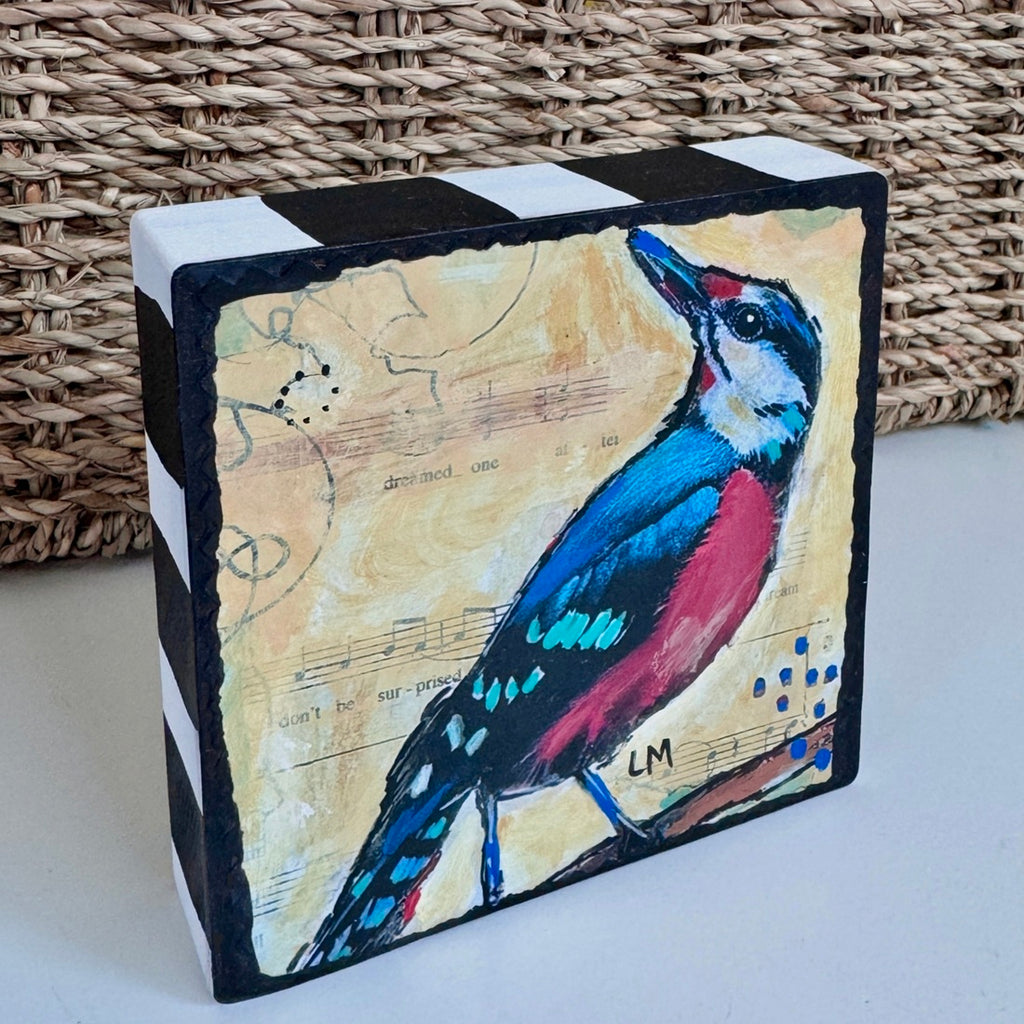 a colorful bird art block painting on a white table with part of a basket showing behind it