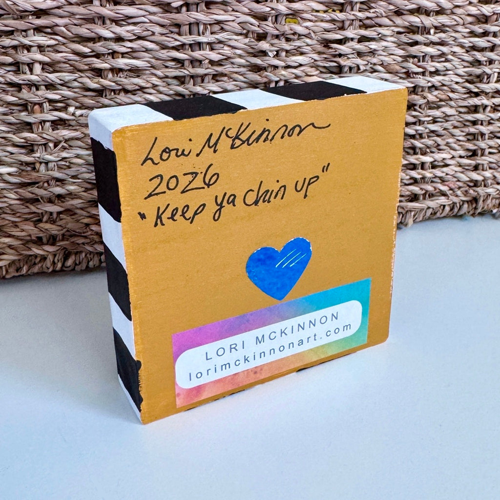the back of a painted wood block that is on a white table with a basket behind it. The block as the artist's name, year and title 'keep ya chin up'