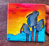 colorful art coaster with three birds at sunset being held by a hand on a dark table