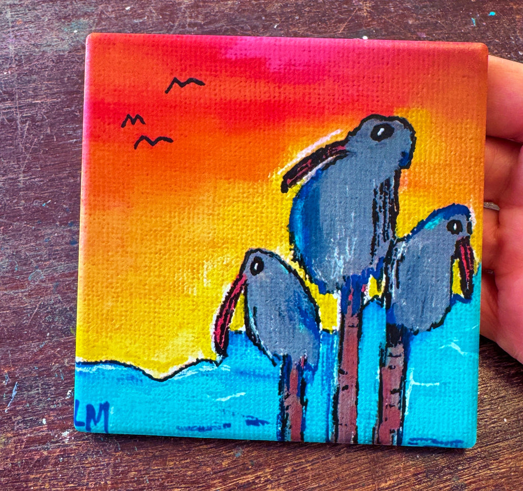 colorful art coaster with three birds at sunset being held by a hand on a dark table