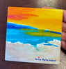 A square coaster featuring a beach scene with a yellow-orange sky, blue water and pale sand. The text 'Anna Maria Island' with a small starfish appears at the bottom.
