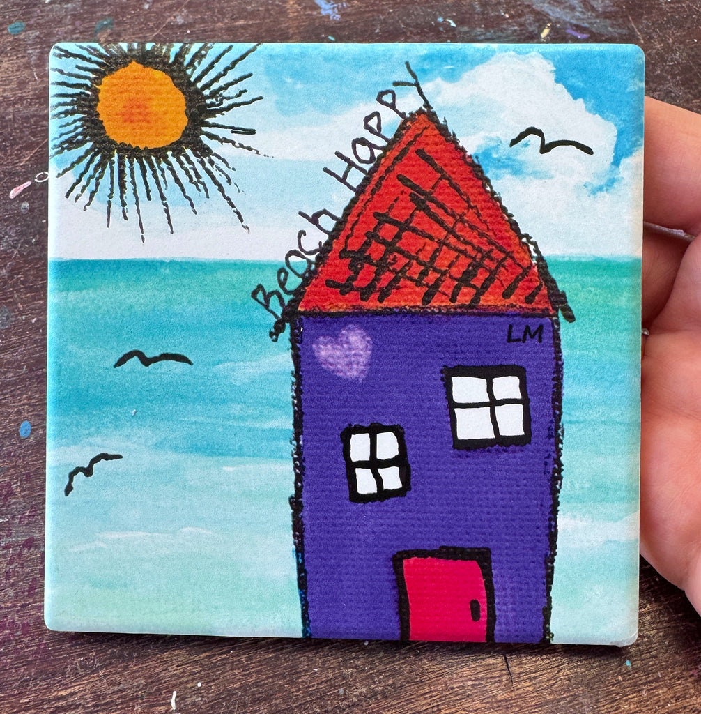 A square coaster with a print of a house by the sea, held by a hand. The painting features a purple house, a bright yellow sun, and a blue ocean under a blue sky