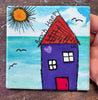 A square coaster with a print of a house by the sea, held by a hand. The painting features a purple house, a bright yellow sun, and a blue ocean under a blue sky