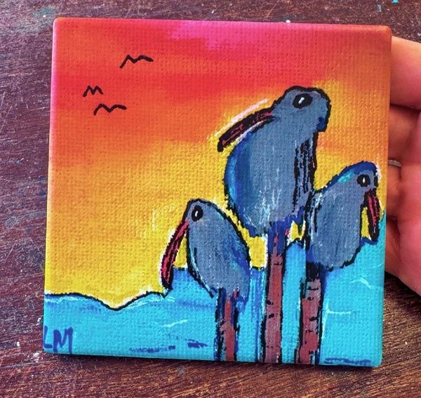 A square coaster featuring three blue birds standing in bright turquoise water against a warm orange and yellow sunset sky, with small black birds in the sky. A hand is holding the coaster on a dark wooden surface.