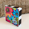 Angled view of a painting art block, showing its colorful flower design and bold black-and-white striped top and side.