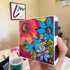 vibrant floral painted art block held in hand, showing its small size and white side painted with black accents