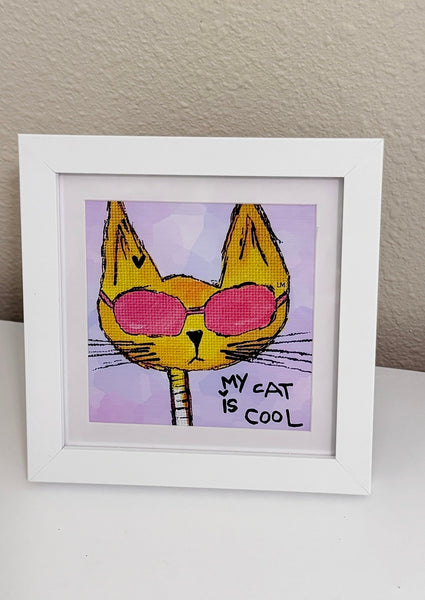 A framed art print featuring a yellow cat wearing pink sunglasses with the text 'MY CAT IS COOL' on a light purple background, displayed in a white frame on a white surface against a beige wall.