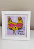 A framed art print featuring a yellow cat wearing pink sunglasses with the text 'MY CAT IS COOL' on a light purple background, displayed in a white frame on a white surface against a beige wall.