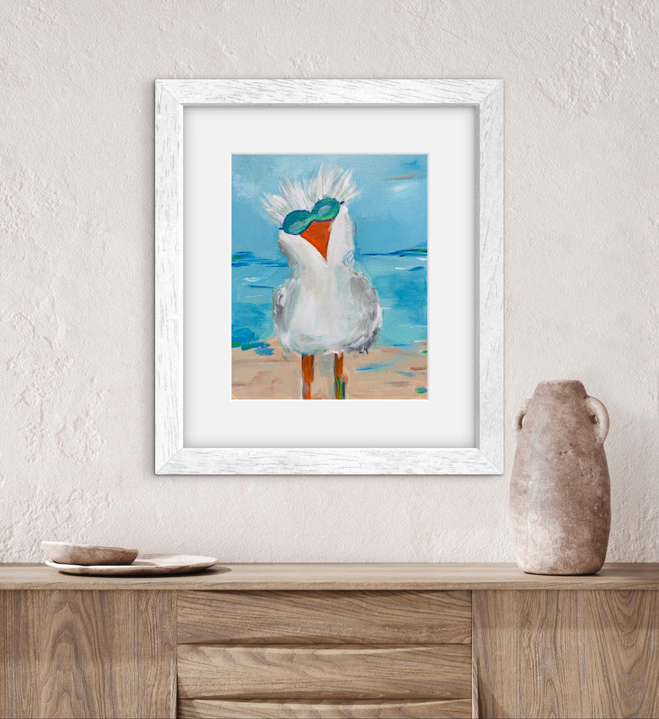 Framed artwork of a seagull on a beach with a vase and plates on a wooden surface below.