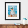 Framed artwork of a seagull with sunglasses on a beach scene, displayed above a wooden surface.