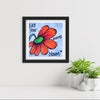 Framed artwork with a red flower and motivational quote on a white wall with plants below.