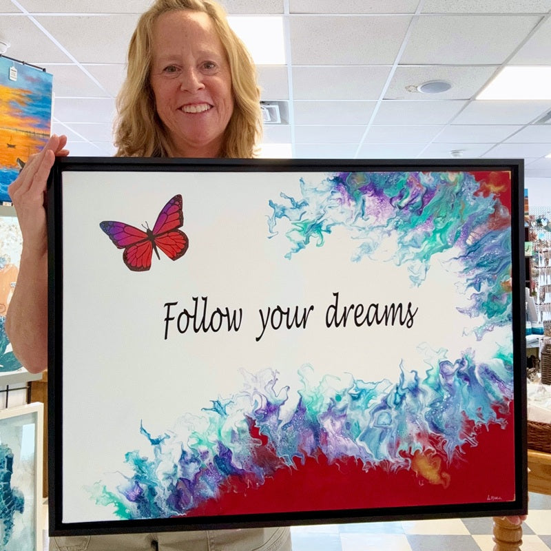 Artist holding a framed artwork with a butterfly and 'Follow your dreams' text in a gallery setting.