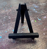 A small black wooden easel photographed on a worn wooden tabletop with paint splatters.