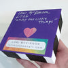 a hand holding a block of wood with purple back showing artist's name, date and signature with a heart