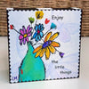 wood block with flowers in a vase and text saying 'enjoy the little things'. There are subtle butterflies in the background of the artwork. 
