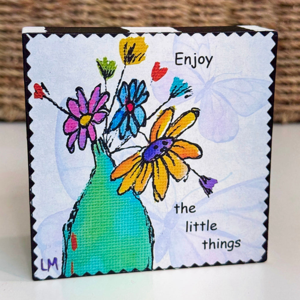 wood block with flowers in a vase and text saying 'enjoy the little things'. There are subtle butterflies in the background of the artwork. 