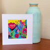 Colorful original floral painting in a white mat, displayed on a wooden tabletop beside a pale turquoise vase.⠋