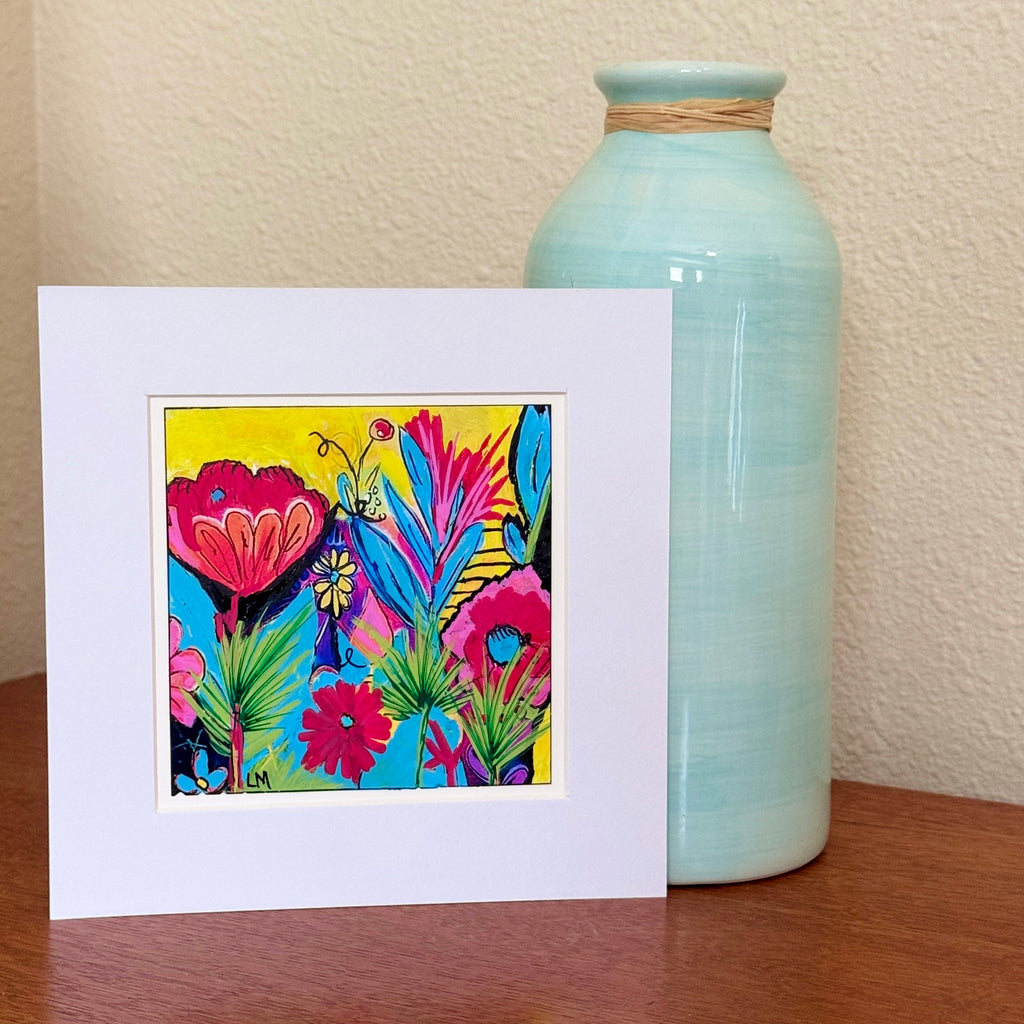 Colorful original floral painting in a white mat, displayed on a wooden tabletop beside a pale turquoise vase.⠋