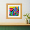 Colorful flower artwork in a natural wood frame on a white wall above a shelf with some vases on it