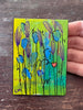 A small painted artwork showing blue flower-like shapes with black stems on a yellow and green background, held by a hand over a dark wooden surface.