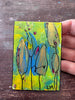 A hand holding a small square abstract painting with green, yellow, and blue brushstrokes, black line details, and small red accents, set against a dark wooden surface.