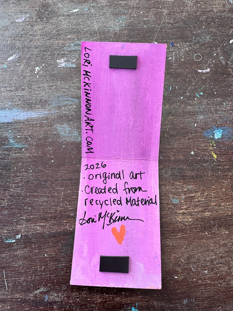 A handmade rectangular art made from recycled cardboard, featuring black magnetic mounting pieces at the top and bottom, handwritten text, and a small orange heart, displayed on a worn wooden surface.