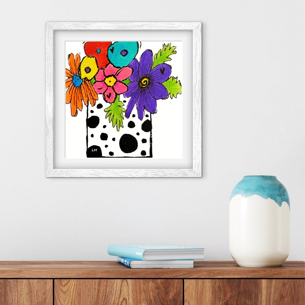 Framed colorful floral artwork on a wall above a wooden shelf with a vase and books.