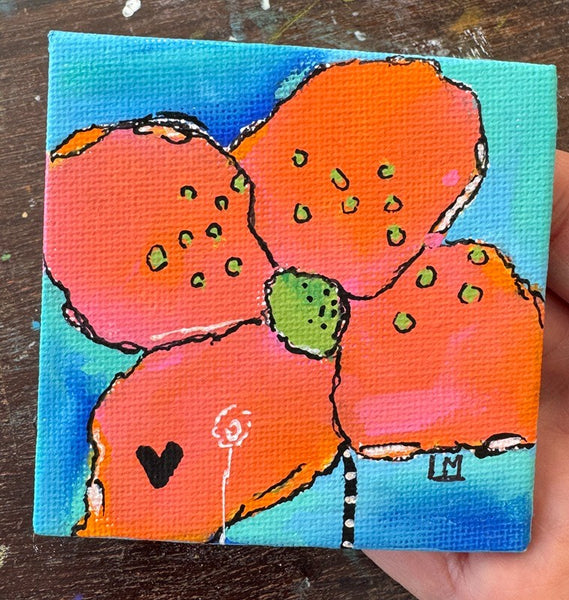 A small square canvas painting of a flower with bright orange petals, a green center, and a blue background, held in a hand over a dark wooden surface.