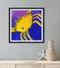 Framed artwork of a yellow crab on a blue background, displayed on a wall with a vase and starfish below.