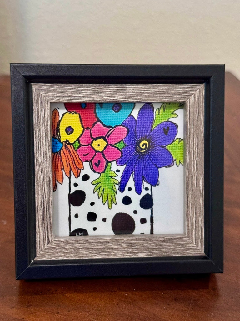 A square framed art print featuring a colorful floral design with a black and white polka dot vase, housed in a black frame with a rustic wood inner frame, resting on a wooden surface.