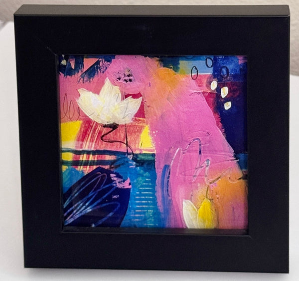 A framed abstract art print featuring a large pink shape, a white flower, and some shades of blue presented in a black square frame against a light background