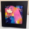 A square black picture frame containing an abstract print with pink, blue, yellow, and white brushstrokes, including a white flower, displayed on a white surface.