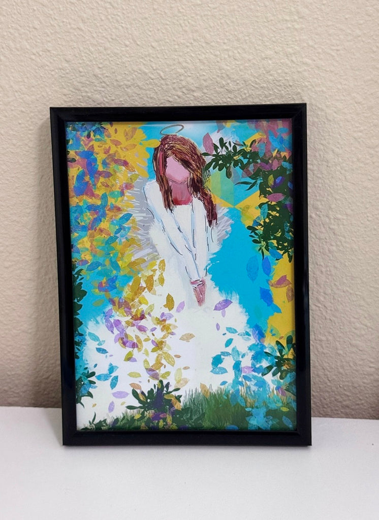 A framed colorful art print of an angel in a white robe standing among vibrant blue, yellow, and green brushstrokes, displayed against a beige wall on a white surface.
