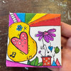 A small square canvas painting held by a hand, featuring a colorful doodle-style design with a yellow guitar, pink heart, purple flower, rainbow stripes, and green leaves on a rustic wooden background.