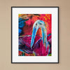 A framed abstract art print featuring a stylized blue bird against a vivid background of pink, red, orange, and blue brushstrokes, displayed on a beige wall.