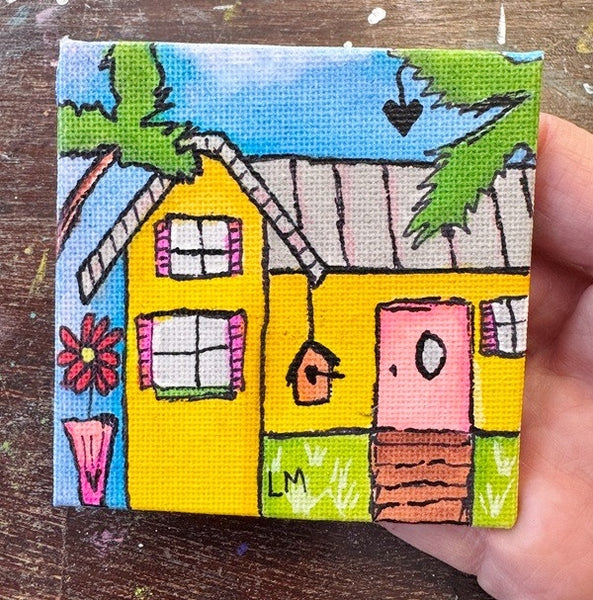 A hand holding a small square canvas painting of a colorful house with a yellow body, pink door and flower pot, green palm trees, and a blue sky, photographed over a wooden surface.