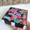 a flower painting on a wood block held by a hand in front of a basket on a white table