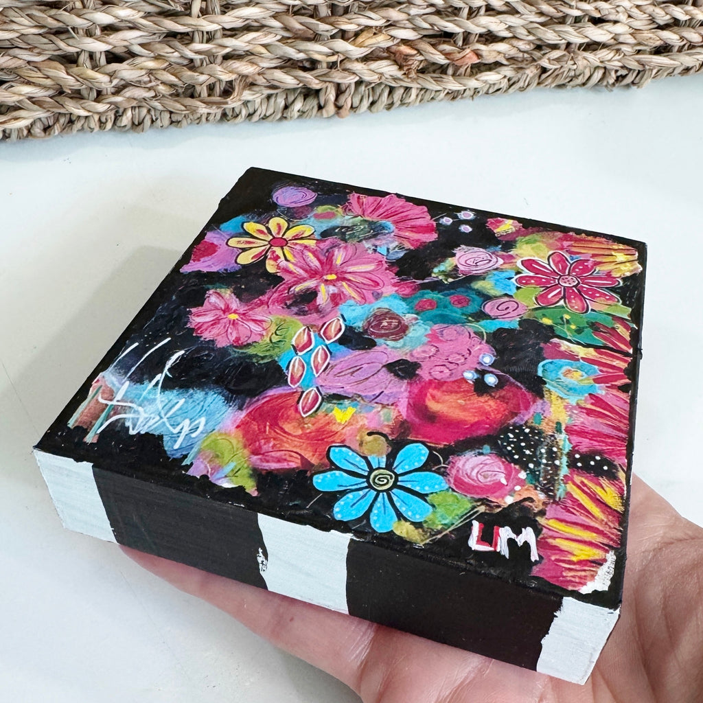 a flower painting on a wood block held by a hand in front of a basket on a white table