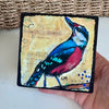 a colorful bird painting held by a hand over a white table with part of a basket showing behind it