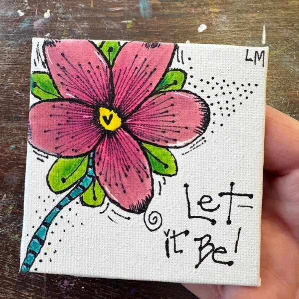 A hand holding a small square canvas painting featuring a pink flower with green leaves, a yellow center, and the handwritten phrase 'Let it Be!' on a rustic wooden background.