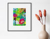 Framed colorful floral artwork on a white wall with a vase of flowers below.
